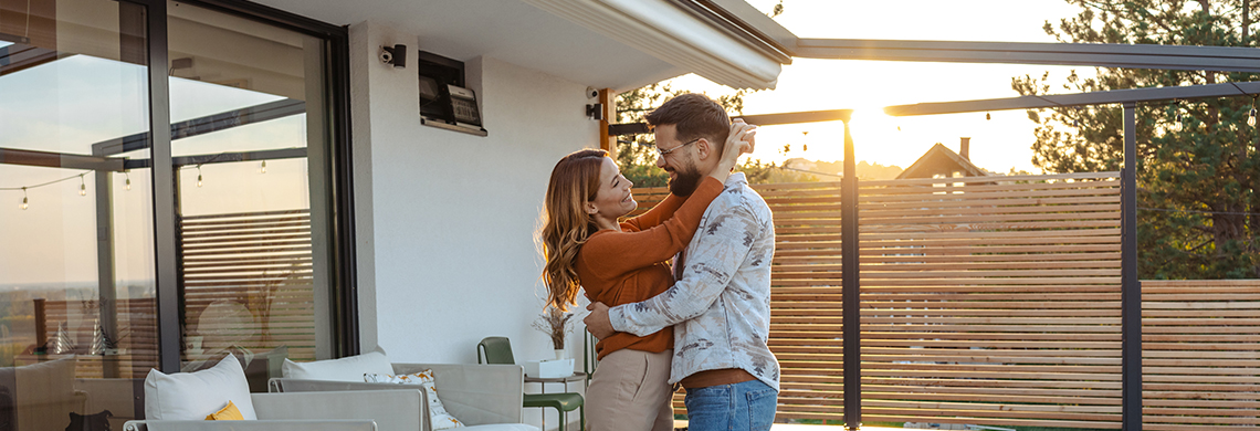 Zwei Personen umarmen sich auf einer modernen Terrasse bei Sonnenuntergang, mit Sitzmöbeln, Tisch, Kissen und Sichtschutzwand.